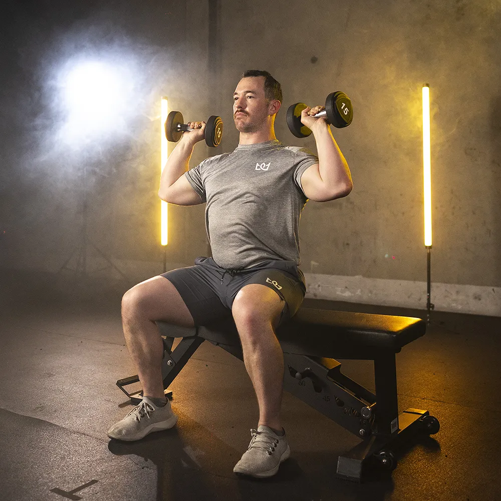 Trainer seated on a bench pressing dumbbells overhead in a studio lit by tall lights.